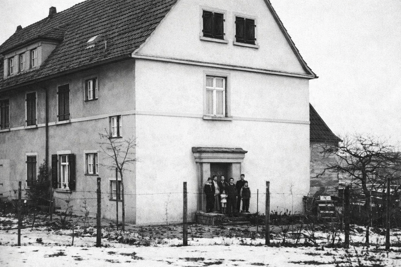 Archivfoto: Die Stirnseite eines weißen Hauses von weiter weg aufgenommen. In der Haustüre stehen mehrere Personen. Privat; Das ehemalige Zollhaus vom Brotdorf; vor der Haustüre stehen Mitglieder der Familie Engel-Berger, um 1936/37
